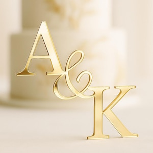 May include: A gold-coloured cake topper displaying the letters "A & K" in a stylish script. The topper is positioned in front of a blurred, two-tiered white cake, suitable for a wedding or celebration.