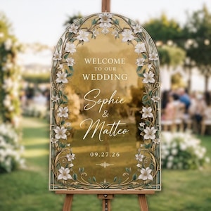 May include: A gold-toned wedding welcome sign with an arched top, decorated with white floral and green foliage. The sign displays "Welcome to our Wedding", the names "Sophie & Matteo", and the date "09.27.26". It is presented on a wooden easel.