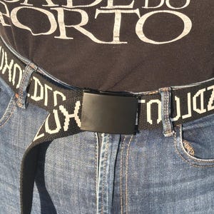 May include: A black fabric belt featuring a repeating white pixelated text pattern and a solid matte black rectangular metal buckle. The belt is threaded through the belt loops of blue denim jeans, worn over a black shirt with partial white text.