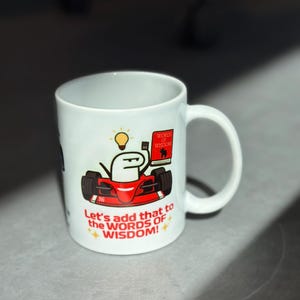 May include: White ceramic mug featuring a cartoon image of a red race car with a stick figure driver holding a book and lightbulb. The mug has the text "Let's add that to the WORDS OF WISDOM!"