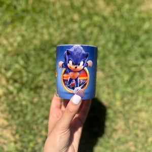 Pode incluir: Uma caneca cilíndrica azul com o personagem Sonic the Hedgehog. A caneca tem uma imagem vibrante de Sonic em ação contra um fundo de pôr do sol. A caneca é segurada contra um fundo de grama verde.