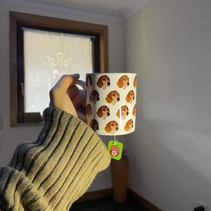 Pode incluir: Uma caneca de cerâmica branca com um padrão repetido de rostos de cães marrons e brancos. A caneca tem um saquinho de chá verde pendurado na alça.