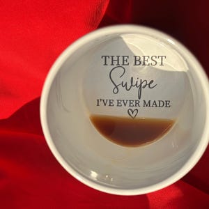 Pode incluir: Caneca de café de cerâmica branca com o texto "THE BEST Swipe I'VE EVER MADE" em escrita preta. A caneca contém uma pequena quantidade de líquido castanho escuro. A caneca é segurada contra um fundo vermelho.