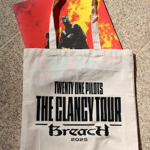 Könnte beinhalten: Eine weiße Canvas-Tragetasche mit dem Text "TWENTY ONE PILOTS THE CLANCY TOUR Breach 2025" in Schwarz. Im Inneren der Tasche befindet sich ein rot-gelber Grafikdruck. Die Tasche hat cremefarbene Griffe.