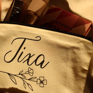 May include: A beige canvas makeup bag with a black zipper and the name "Tixa" written in black cursive script. A floral design is below the name. The bag is partially open and contains makeup products.