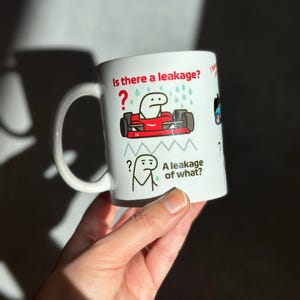 Può includere: Tazza in ceramica bianca con manico. Presenta un design a cartoni animati con il testo "Is there a leakage?" e "A leakage of what?" insieme a illustrazioni di un'auto da corsa e un omino stilizzato.