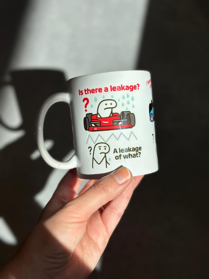 May include: White ceramic mug with a handle featuring a humorous cartoon design. The mug has the text "Is there a leakage?" and "A leakage of what?" along with cartoon illustrations of a race car and a stick figure.
