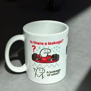 May include: White ceramic mug with a handle, featuring a cartoon design. The design includes a red race car with a stick figure driver and the text "Is there a leakage?" and "A leakage of what?"