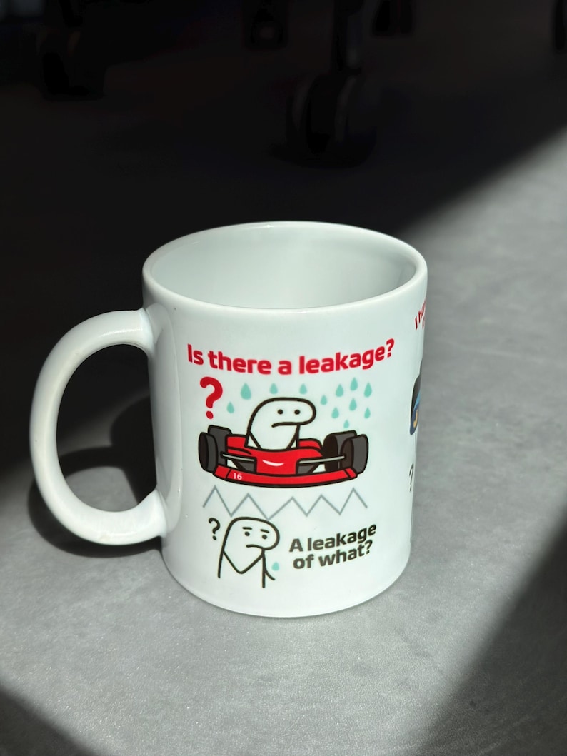 May include: White ceramic mug with a handle, featuring a cartoon design. The design includes a red race car with a stick figure driver and the text "Is there a leakage?" and "A leakage of what?"
