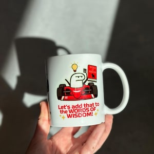 May include: White ceramic mug featuring a cartoon character in a red race car, holding a book labeled "Words of Wisdom." The mug also has the text "Let's add that to the WORDS OF WISDOM!" with star accents.
