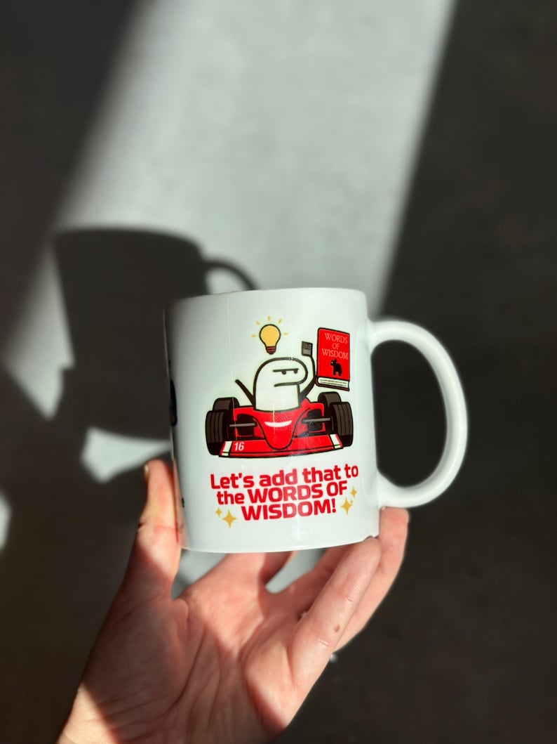 May include: White ceramic mug featuring a cartoon character in a red race car, holding a book labeled "Words of Wisdom." The mug also has the text "Let's add that to the WORDS OF WISDOM!" with star accents.