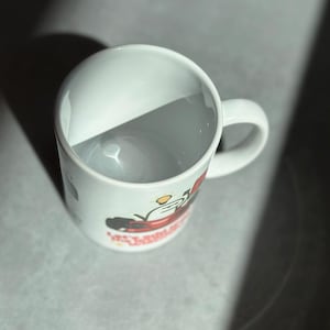 May include: White ceramic mug with a curved handle. The mug features a colorful graphic design on the side. The mug is empty and sits on a gray surface, with a shadow cast across it.