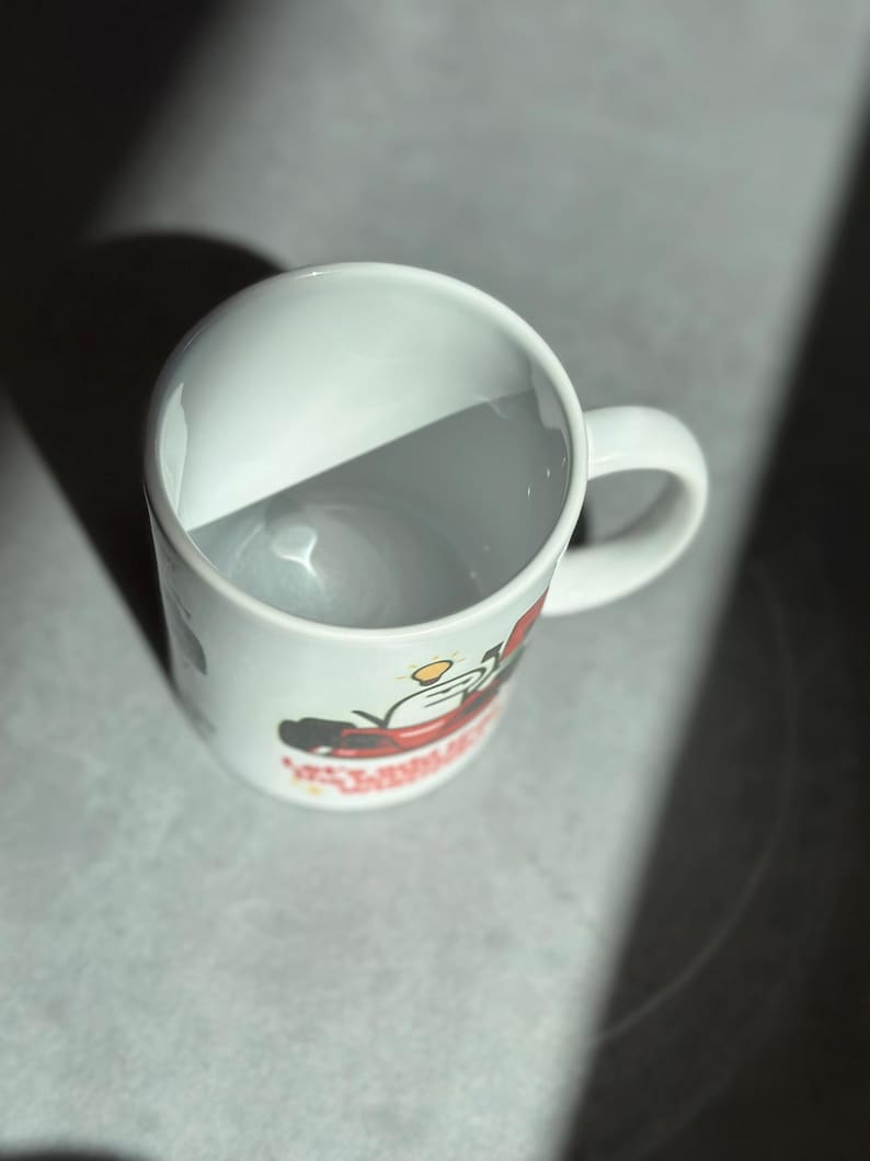 May include: White ceramic mug with a curved handle. The mug features a colorful graphic design on the side. The mug is empty and sits on a gray surface, with a shadow cast across it.