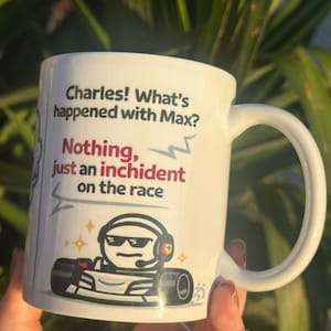 Pode incluir: Caneca de cerâmica branca com uma alça curva. A caneca apresenta o texto "Charles! O que aconteceu com o Max? Nada, apenas um incidente na corrida". Um desenho animado de um piloto de corrida também é retratado.