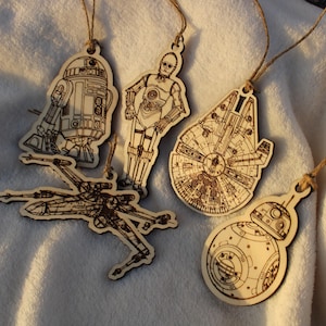May include: Five wooden ornaments featuring characters from Star Wars. The ornaments are R2-D2, C-3PO, a TIE fighter, a Millennium Falcon, and BB-8.