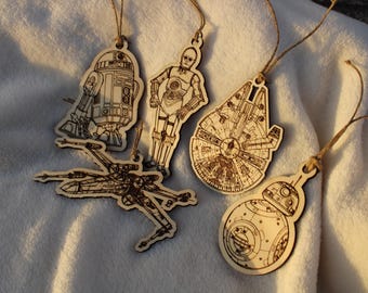 Könnte beinhalten: Fünf Holzornamente mit Figuren aus Star Wars. Die Ornamente sind R2-D2, C-3PO, ein TIE-Jäger, ein Millennium Falcon und BB-8.