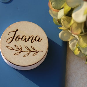 Op de afbeelding: Een ronde houten telefoonhouder met de naam "Joana" gegraveerd in cursief schrift. Onder de naam bevindt zich een ontwerp met een bladertak.