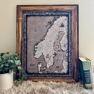Puede incluir: Mapa enmarcado de Escandinavia, con el texto "SKANDINAVIA AD 900". El mapa es en tonos grises y negros, con detalles intrincados de la tierra y el mar. El marco es de madera con un acabado oscuro.