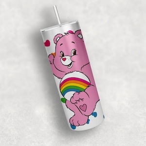 Peut inclure: Un gobelet en acier inoxydable blanc avec une paille, sur lequel est représenté un Care Bear rose avec un arc-en-ciel sur le ventre et des cœurs sur les pieds.