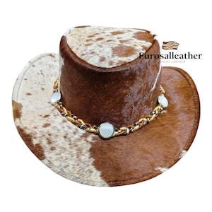 May include: Cowboy hat with a brown and white spotted pattern. The hat features a brown crown and brim with a decorative chain and three white stones. The hat is made of leather and has the text "Eurosalleather".