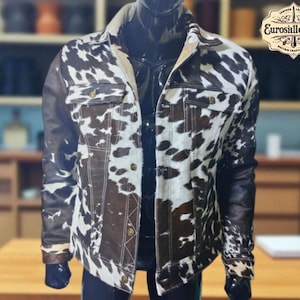 Könnte beinhalten: Jacke mit Kuhfellmuster mit schwarzen Lederärmeln und Kragen. Die Jacke hat eine Knopfleiste vorne, zwei Brusttaschen und einen beigefarbenen Kragen. Die Jacke wird auf einer Schaufensterpuppe präsentiert. Das Eurosilleather-Logo ist sichtbar.