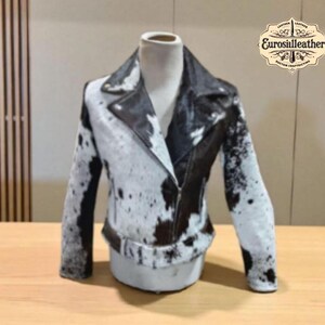Könnte beinhalten: Schwarz-weiße Lederjacke aus Kuhfell mit Reverskragen und Reißverschluss. Die Jacke wird auf einer Schaufensterpuppe präsentiert und weist ein einzigartiges Fleckenmuster auf. Die Jacke hat lange Ärmel und einen Gürtel.