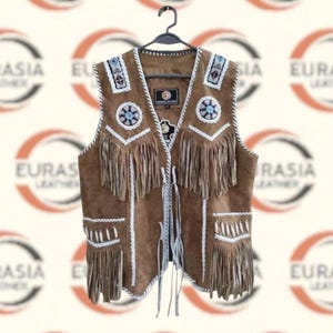 Puede incluir: Chaleco de ante marrón con ribete blanco, flecos y abalorios decorativos. El chaleco tiene cuello en V y cierre de lazo. El chaleco está colgado de una percha negra. El chaleco está decorado con detalles blancos y azules.
