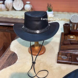 May include: A black leather hat with a wide brim and a decorative band featuring silver accents. The hat is displayed on a wooden stand with a black chin strap. The hat is a western style.