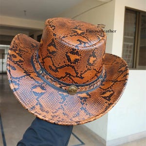 Puede incluir: Un sombrero de vaquero marrón y negro con estampado de piel de serpiente. El sombrero presenta un ala ancha, una banda decorativa con remaches y un emblema circular. El texto "Eurosalleather" es visible.