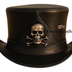 Könnte beinhalten: Schwarzer Lederzylinder mit silbernem Totenkopf- und gekreuzten Knochen-Emblem auf der Vorderseite. Der Hut hat eine breite Krempe und eine hohe, zylindrische Krone. Das Emblem befindet sich auf einem dreieckigen Aufnäher.
