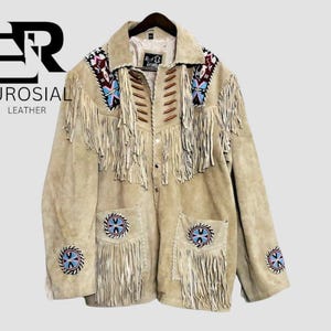May include: A beige suede leather jacket featuring long fringe detailing across the chest and pockets. The jacket includes colorful beaded circular patches on the sleeves and pockets, brown wooden bead accents, and the text EUROSIAL LEATHER.