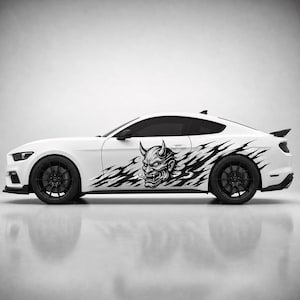 Puede incluir: Un coche deportivo blanco con un gran adhesivo gráfico negro en la puerta lateral. El diseño muestra una máscara Hannya detallada con cuernos, rodeada de patrones negros estilizados en forma de llama que se extienden por la carrocería.