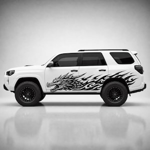 Puede incluir: Un SUV blanco con una gran calcomanía gráfica negra en el panel lateral. La calcomanía representa una cabeza de dragón detallada con la boca abierta, que se transforma en patrones de llamas estilizados que se extienden hacia la parte trasera.