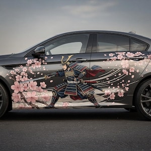 May include: Dark grey car featuring a detailed graphic of a samurai warrior in traditional armour, wielding a sword. Pink cherry blossom flowers are painted on the car's side, creating a striking contrast. The car is parked on a paved surface.