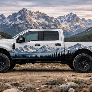 Op de afbeelding: Een witte pick-up truck met een berglandschapontwerp op de zijkant en achterkant. De truck heeft zwarte wielen en banden en staat geparkeerd op een rotsachtig oppervlak met bergen op de achtergrond.