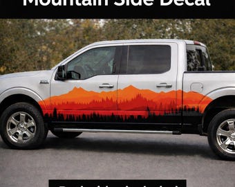 May include: A silver pickup truck with a mountain side decal. The decal features a layered mountain range design in shades of orange and red, with a black forest silhouette at the base. The text "Mountain Side Decal" is at the top.