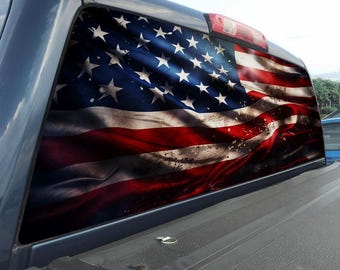 May include: A perforated vinyl window decal applied to the rear glass of a pickup truck, featuring a high-resolution, weathered graphic of the American flag with red, white, and blue colors and a distressed, textured appearance.