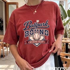 May include: A brick red short-sleeve t-shirt featuring a graphic print with the text Ballpark Bound and Weekend Ready. The design includes a baseball illustration centered within a home plate shape, rendered in navy blue, white, and red colors.