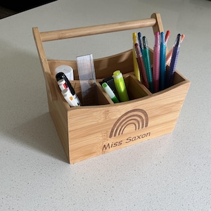 May include: A light brown bamboo desk organiser with a handle, featuring a laser-engraved rainbow design and the text "Miss Saxon" on the front. The organiser has multiple compartments filled with various coloured markers, pens, and a clear ruler.