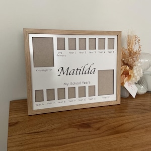 May include: A light brown wooden framed photo display for school years, with spaces for photos from Kindergarten to Year 12. The display is personalised with the name "Matilda" and sits on a wooden surface.