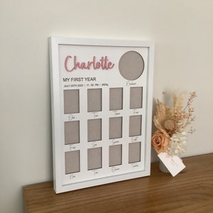 May include: White picture frame with a pink inscription that reads "Charlotte My First Year" and a grid of 12 photo spaces. The frame is on a wooden surface with a small bouquet of dried flowers in a white vase.