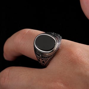 May include: A silver ring with an oval, black stone set in a decorative band. The ring has intricate patterns and is worn on a finger. The background is a solid black.