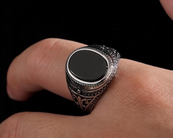 May include: A silver-colored ring with an oval, black stone set in a decorative band. The ring features intricate patterns and is worn on a finger. The background is a solid black.