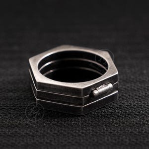 Peut inclure: Une bague géométrique argentée avec un design hexagonal. La bague présente une construction en couches avec un petit fermoir rectangulaire. La bague est posée sur un fond sombre, soulignant sa finition métallique.