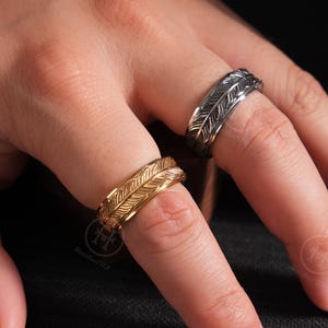 May include: Two feather-design rings, one gold-toned and one silver-toned, are displayed on a hand. The rings have detailed feather patterns. The background is dark, emphasizing the jewelry.