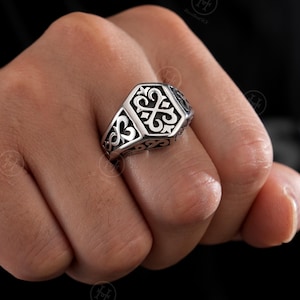 May include: A silver ring with an ornate, hexagonal face. The ring features a detailed, symmetrical pattern of swirling lines and shapes. The ring is worn on a finger, showcasing its intricate design and craftsmanship. The ring's design is a classic style.