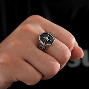 Può includere: Un anello color argento con un design a stella su sfondo scuro. L'anello è indossato su un dito, mostrando la sua dettagliata lavorazione e il motivo geometrico. Il design dell'anello presenta un emblema a stella.