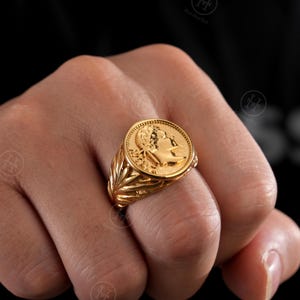 May include: A gold coin ring featuring a detailed profile on the coin's face. The ring has a textured band with a leaf design. The ring is displayed on a hand against a dark background.