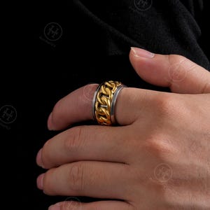 Peut inclure: Une bague à maillons dorés et argentés. La bague présente un motif de chaîne dorée au centre, encadré de bandes argentées. La bague est portée au doigt, mettant en valeur son design et ses couleurs métalliques.