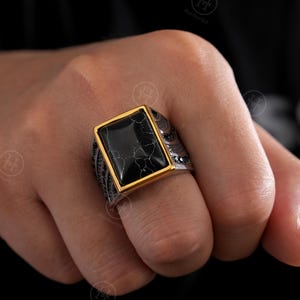 May include: A silver ring with a rectangular black stone set in a gold-colored bezel. The ring has intricate detailing on the band. The black stone has a cracked, marbled appearance.